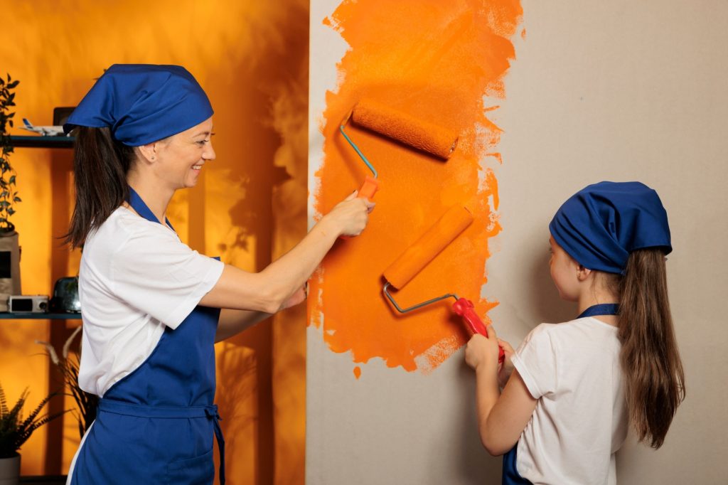 Mother and child using orange paint on house walls to renovate apartment room, having fun with housework redecoration. Woman with little kid painting with paintbrush and roller