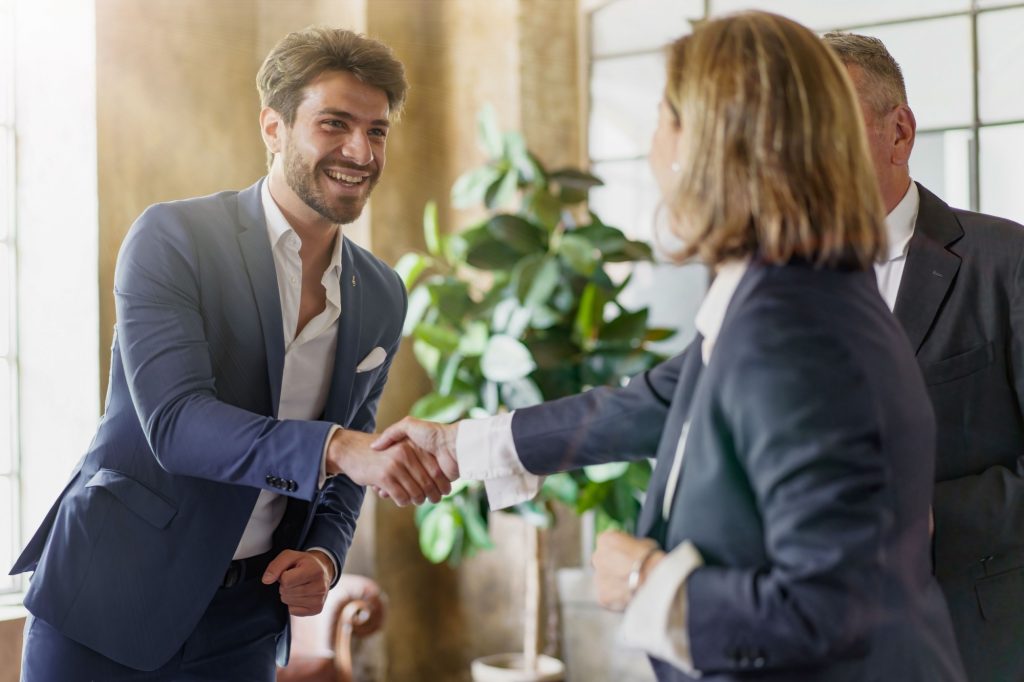 Smiling businessman shaking hands with colleagues in modern office. Professional greeting, successful partnership. Corporate environment, positive business interaction, teamwork concept.