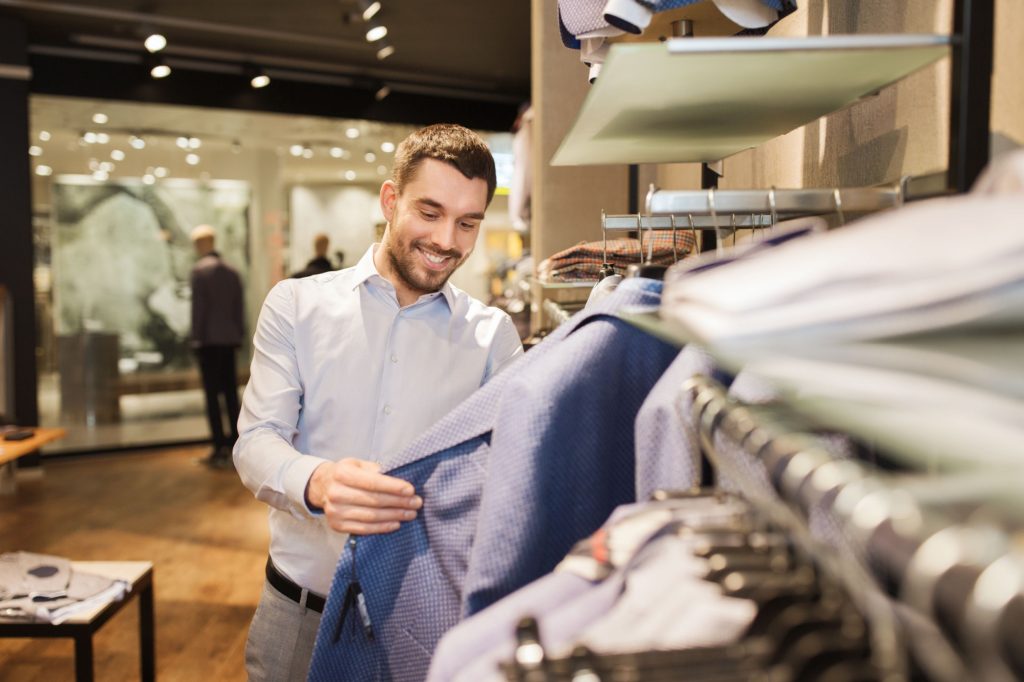 Sale, shopping, fashion, style and people concept - happy young man in shirt choosing jacket in mall or clothing store