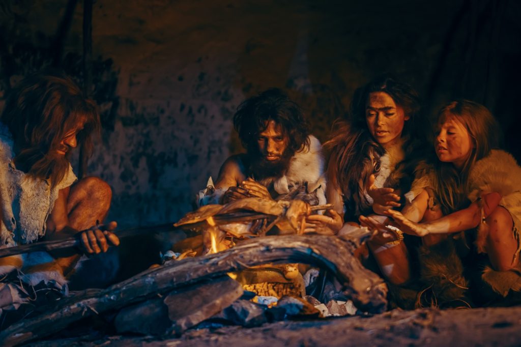 Neanderthal or Homo Sapiens Family Cooking Animal Meat over Bonfire and then Eating it. Tribe of Prehistoric Hunter-Gatherers Wearing Animal Skins Grilling and Eating Meat in Cave at Night