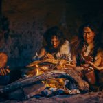 Neanderthal or Homo Sapiens Family Cooking Animal Meat over Bonfire and then Eating it. Tribe of Prehistoric Hunter-Gatherers Wearing Animal Skins Grilling and Eating Meat in Cave at Night