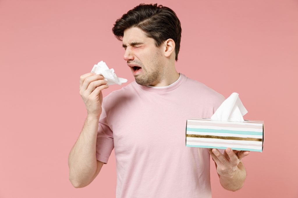 Sick unhealthy ill allergic man has red watery eyes runny stuffy sore nose suffer from allergy trigger symptoms hay fever hold paper napkin handkerchief isolated on pastel pink color background studio