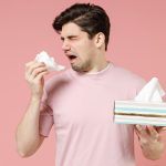 Sick unhealthy ill allergic man has red watery eyes runny stuffy sore nose suffer from allergy trigger symptoms hay fever hold paper napkin handkerchief isolated on pastel pink color background studio