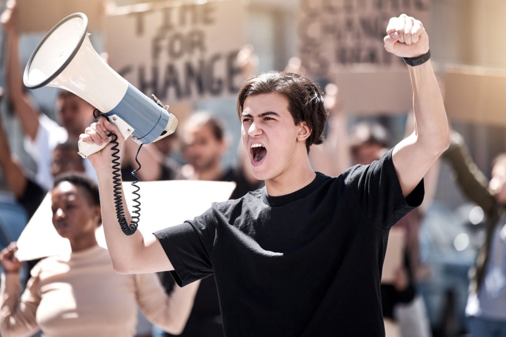 Frustrated man, megaphone and protest with crowd for human rights, equality or change in society or city. Angry male person or activist screaming or yelling on loudspeaker in street riot or rally