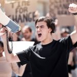 Frustrated man, megaphone and protest with crowd for human rights, equality or change in society or city. Angry male person or activist screaming or yelling on loudspeaker in street riot or rally