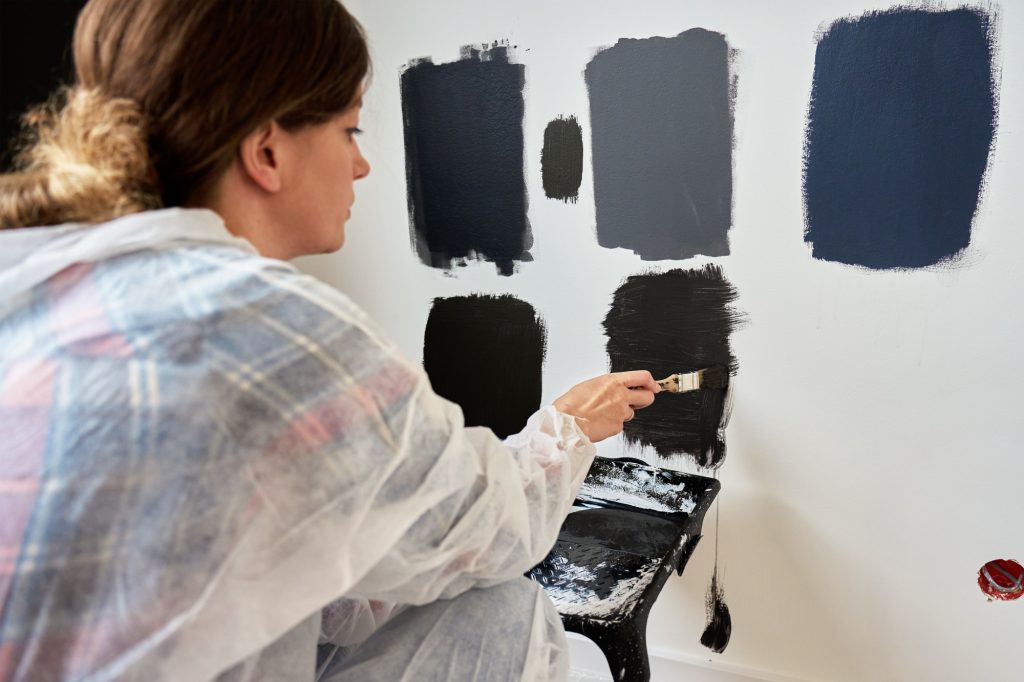 Woman in protective wear applies different dark paint swatches on white wall using brush. Female painter testing black and dark gray paint samples during interior renovation project in modern home