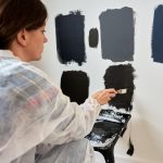 Woman in protective wear applies different dark paint swatches on white wall using brush. Female painter testing black and dark gray paint samples during interior renovation project in modern home