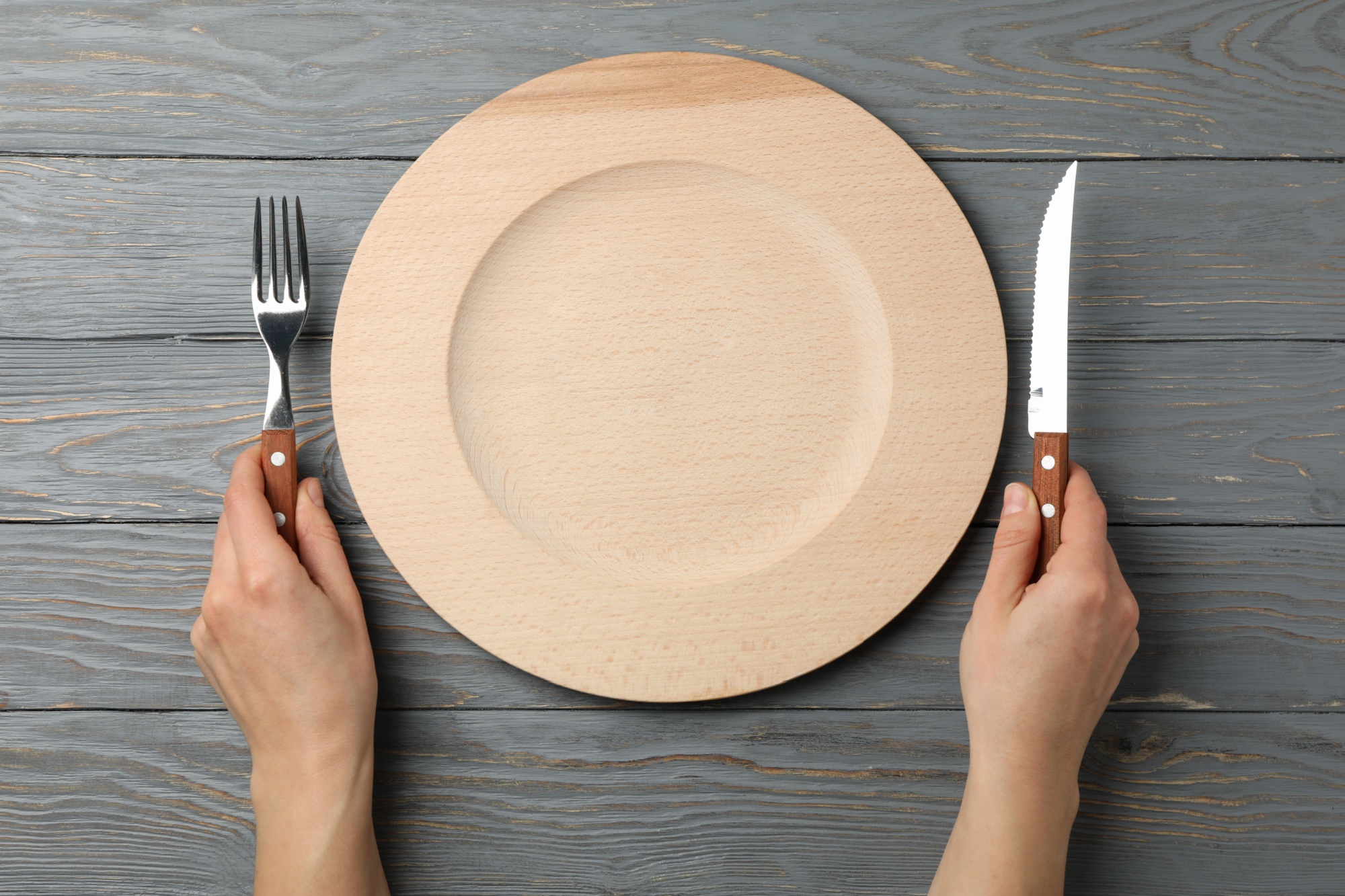 Female hands hold fork and knife on wooden background with plate, top view