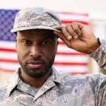 Portrait of confident male african american army soldier wearing cap and uniform against flag. patriotism and identity, unaltered.