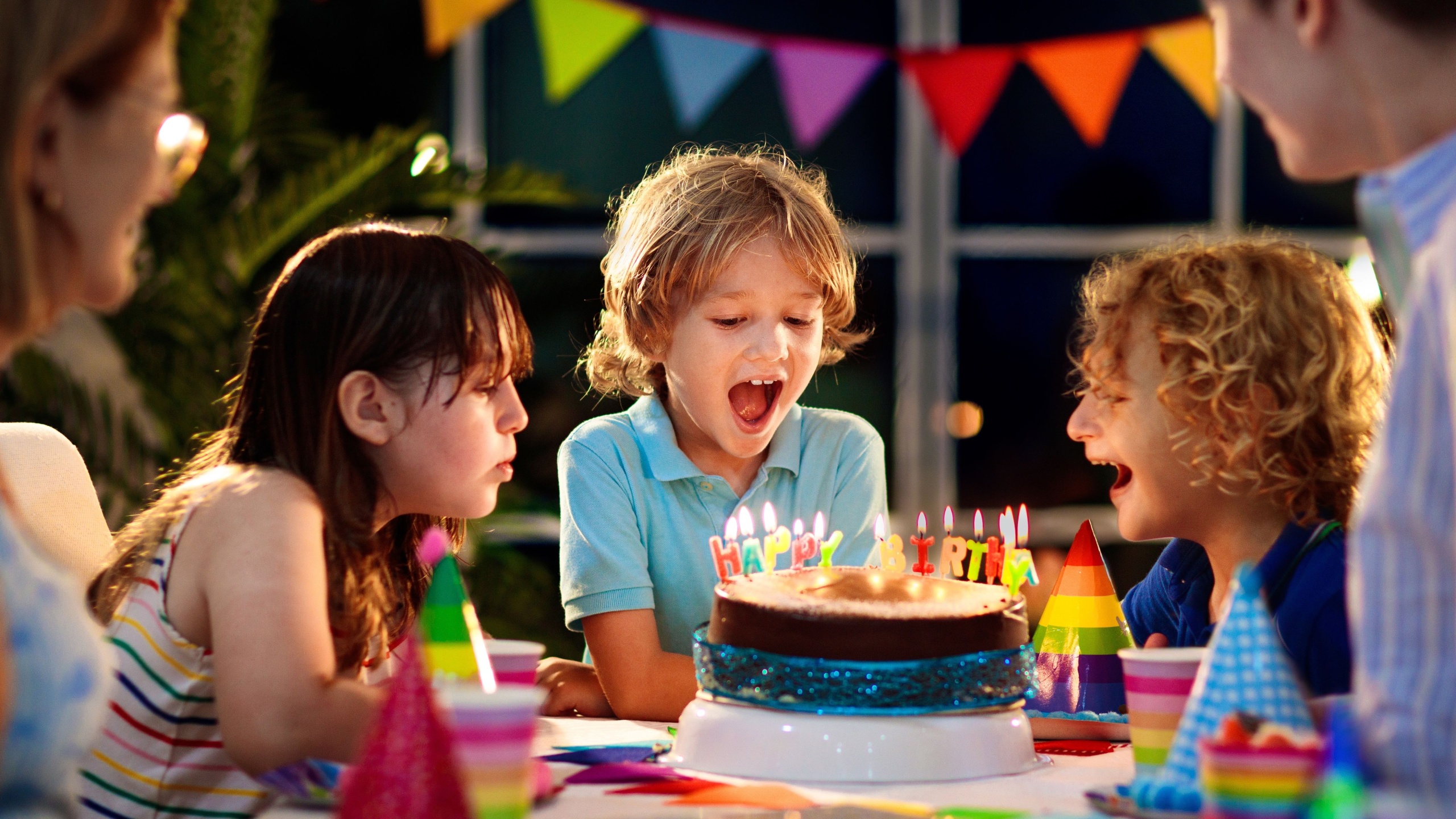 Kids birthday party. Child blowing candles on cake and opening presents. Pastel rainbow theme celebration. Family celebrating at home. Boy opening gifts, eating cakes. Sweets for children.