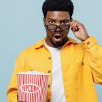 Young shocked amazed scared man of African American ethnicity take off 3d glasses watch movie film hold bucket of popcorn isolated on plain blue background People emotions in cinema lifestyle concept