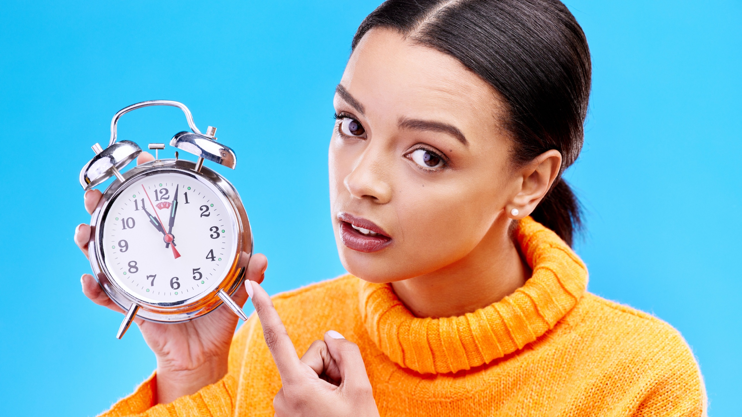 Woman, annoyed and point at alarm clock in portrait for warning by blue background in studio. Gen z girl, student and model with watch, time management and schedule with angry face to start morning