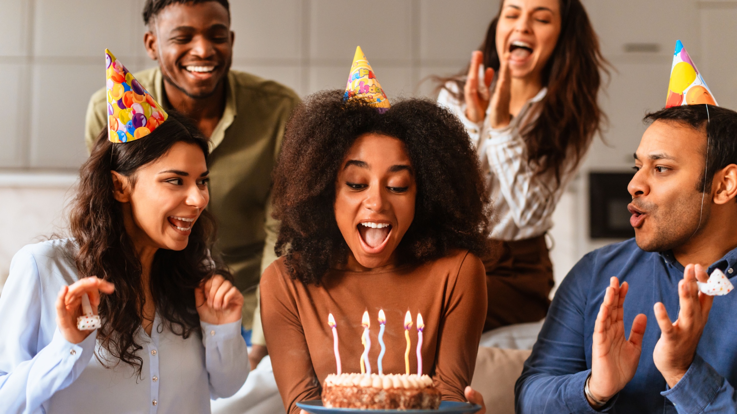 Multiracial friends surround a woman holding a birthday cake, blowing out candles in a moment of excitement together