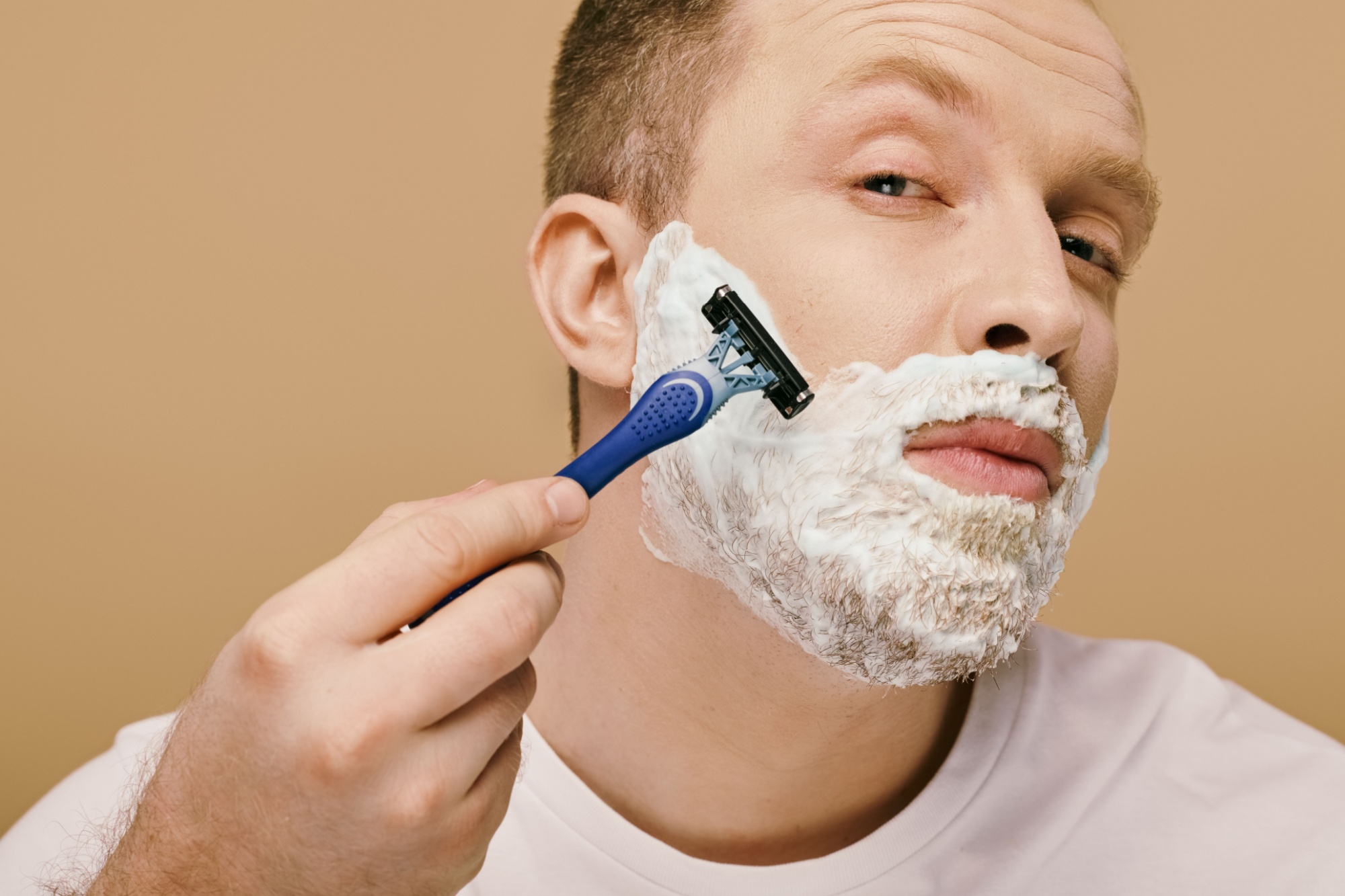 A man in casual attire carefully shaves his face with a razor.