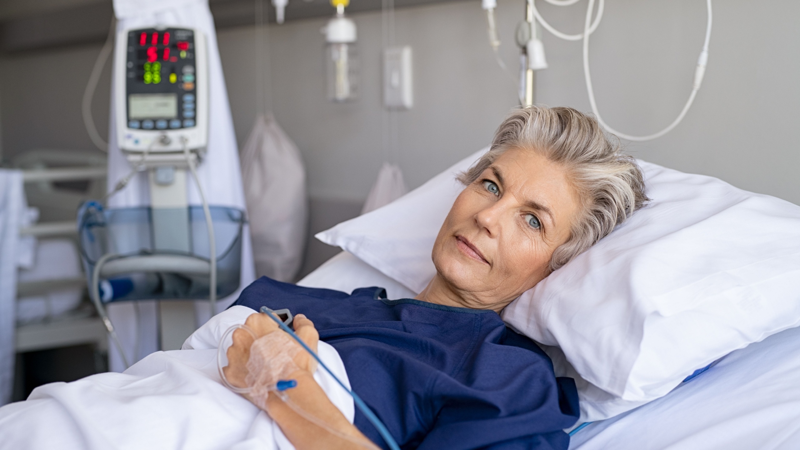 Mature woman recovering from illness while lying on hospital bed with iv drip in hand. Portrait of old hospitalized patient recovering after surgery. Happy senior woman lying on bed in hospital ward.