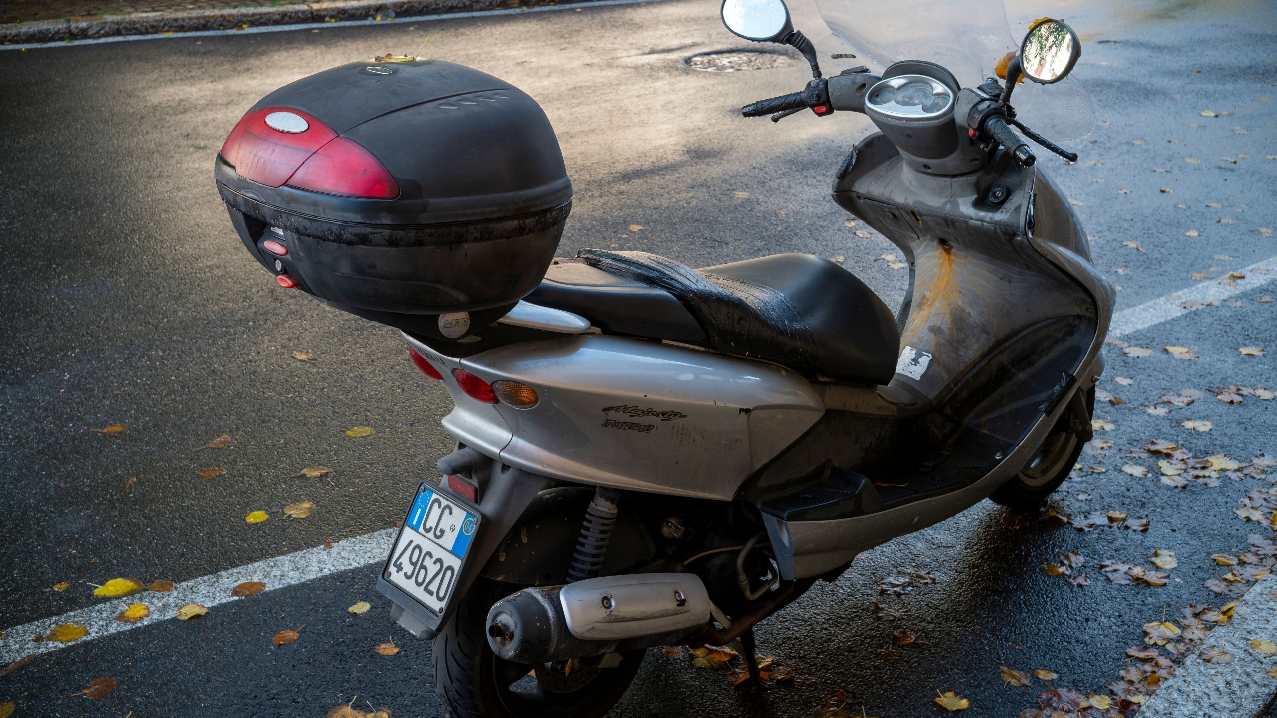 Cremona, Lombardy, Italy October 15th 2024 An old, used silver scooter with a black trunk is parked on the side of a city street in autumn