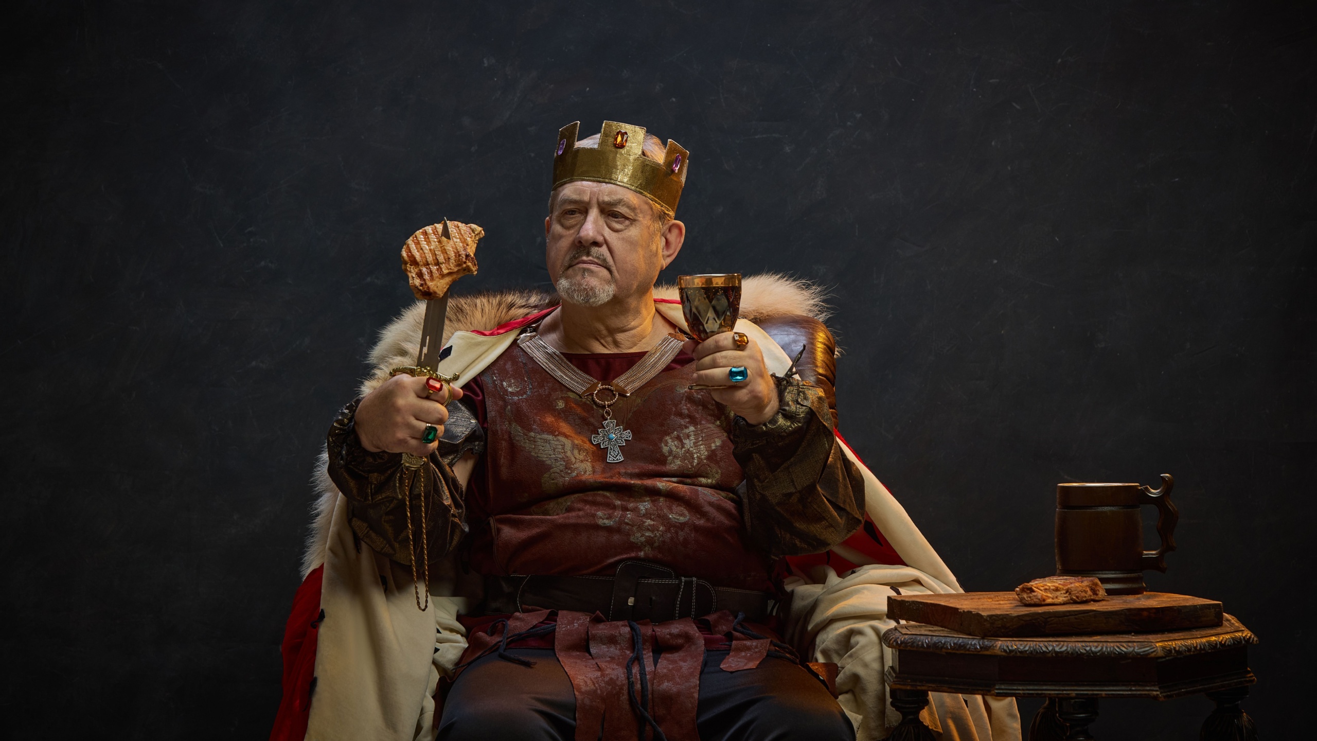 Senior man, medieval king in period attire sitting on throne with meat on sword and glass of wine against black studio background. Concept of history, royalty, power, fantasy, leadership, celebration