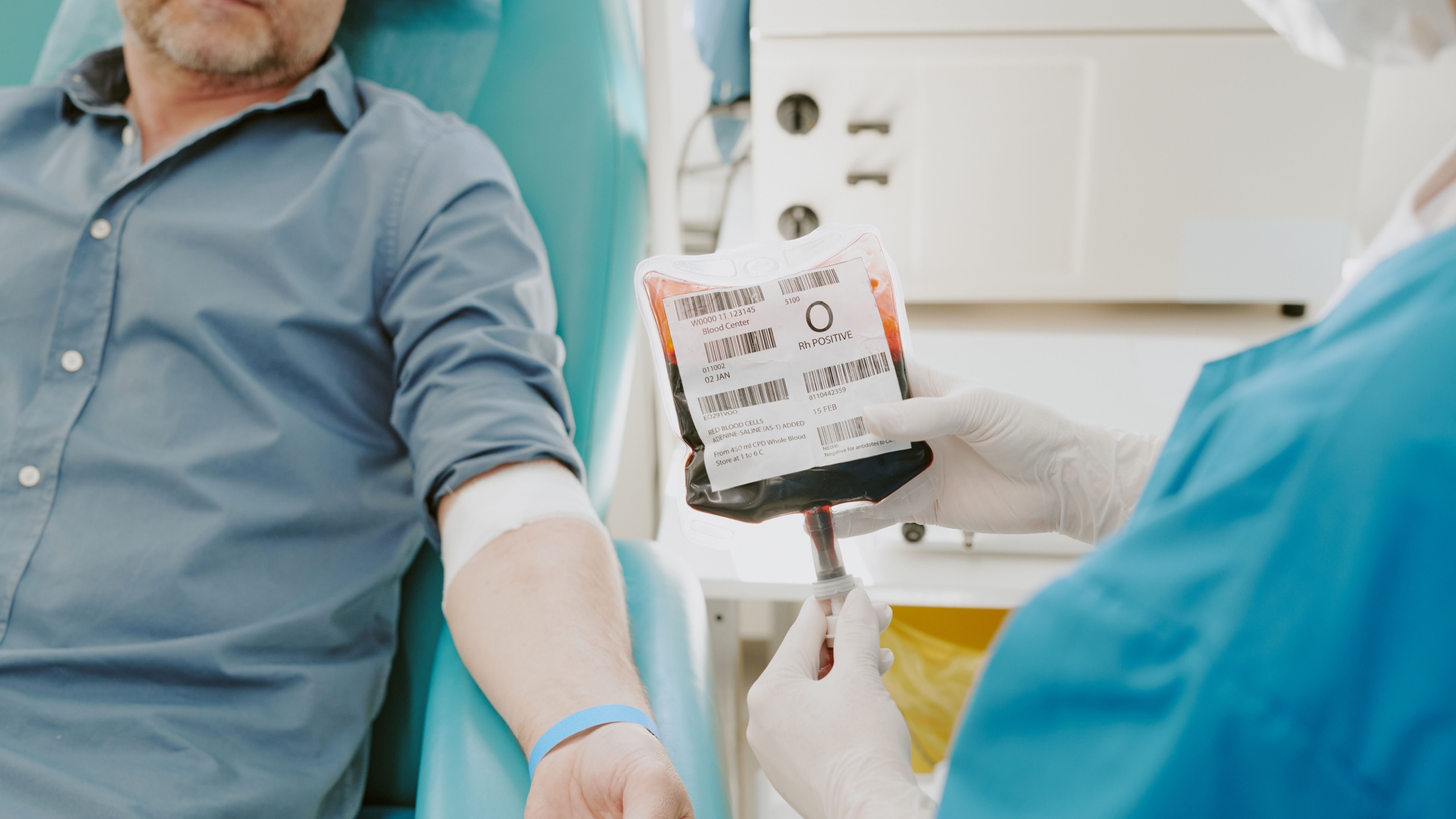 Middle aged Caucasian man donating blood while medical worker holding blood collection bag and monitoring process in clinical setting, visible blood donation equipment and patient arm