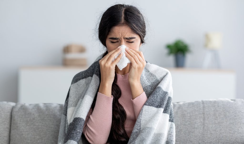 Unhappy sad young indian female in plaid suffering from fever and flu on sofa, blowing nose in napkin in living room interior. Covid-19 lockdown, treatment of illness, cold and runny, copy space