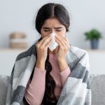 Unhappy sad young indian female in plaid suffering from fever and flu on sofa, blowing nose in napkin in living room interior. Covid-19 lockdown, treatment of illness, cold and runny, copy space