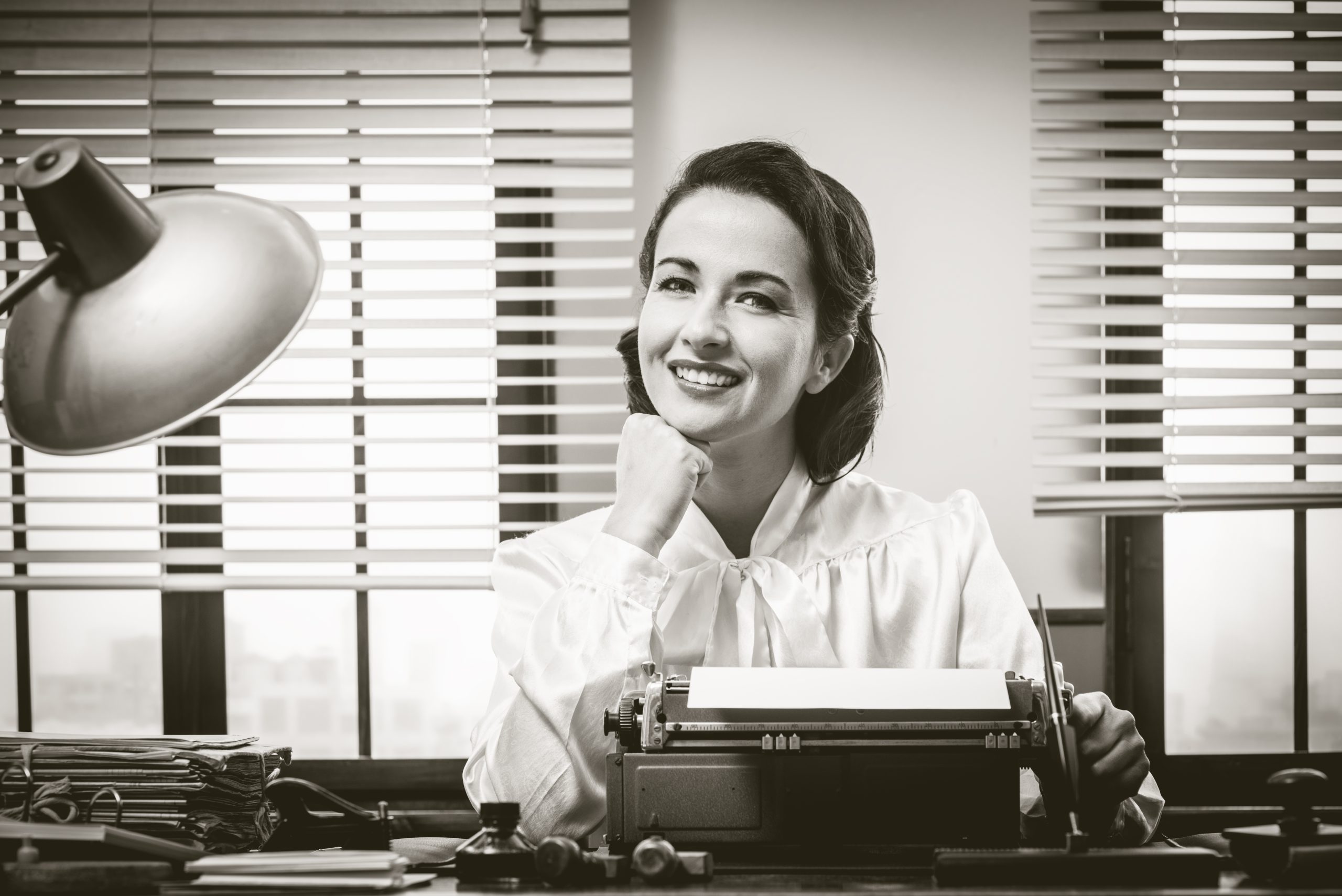 1950s style secretary working at office desk and smiling with hand on chin