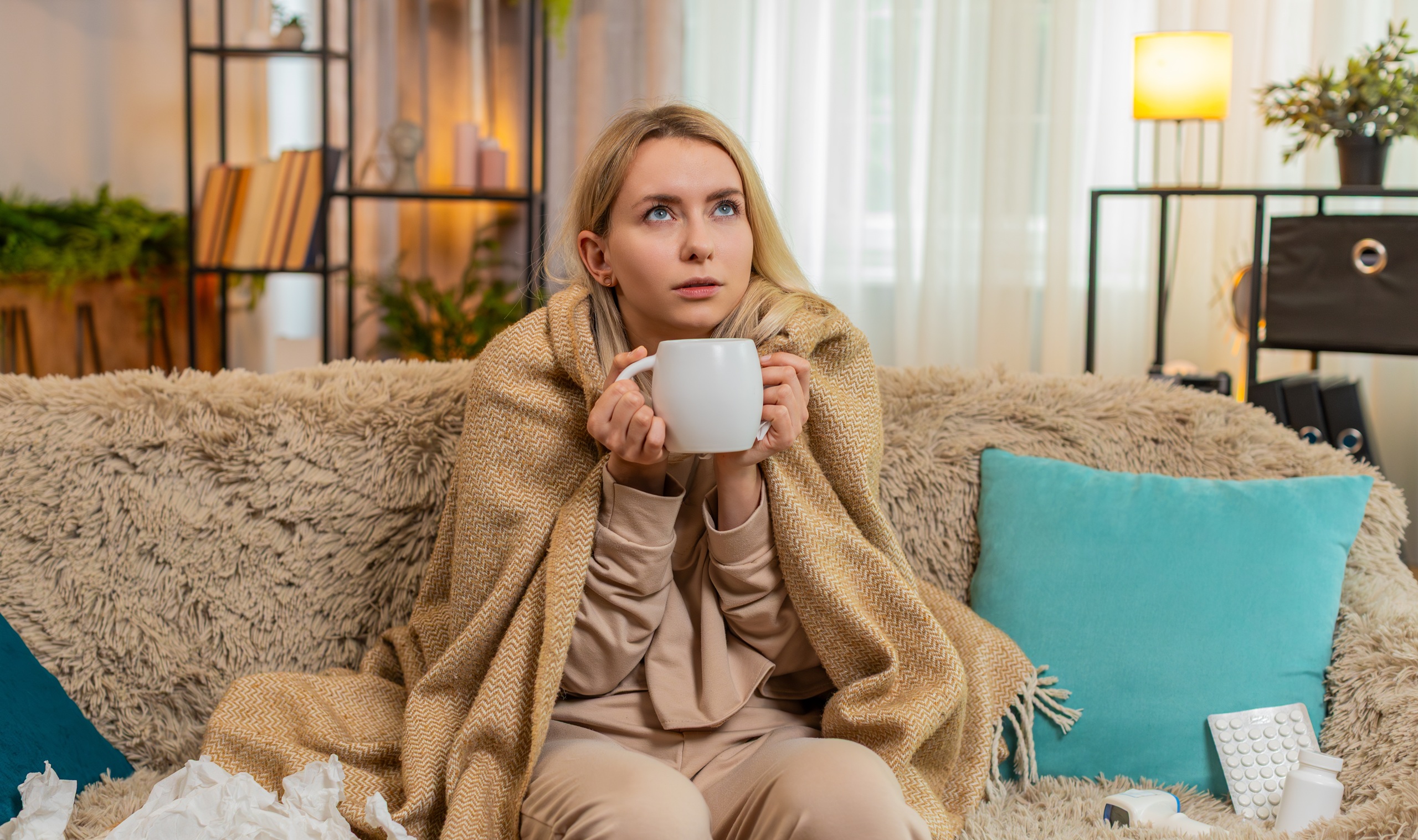 Young ill adult woman sits on home sofa under blanket, holding cup of hot tea and warming hands, sniffling gently. Blonde girl looks tired and sick, reacting to cold symptoms with calm expression.
